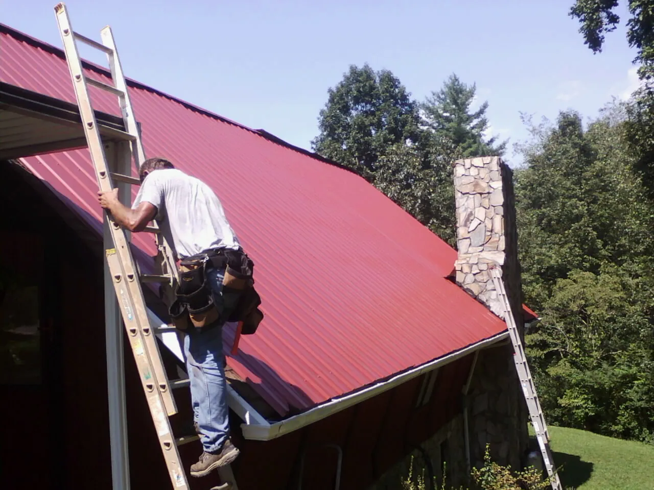 Roofer on a ladder beside a red metal roof and stone chimney — Soffit & Fascia Repair by Roofing Brothers in South Lancaster